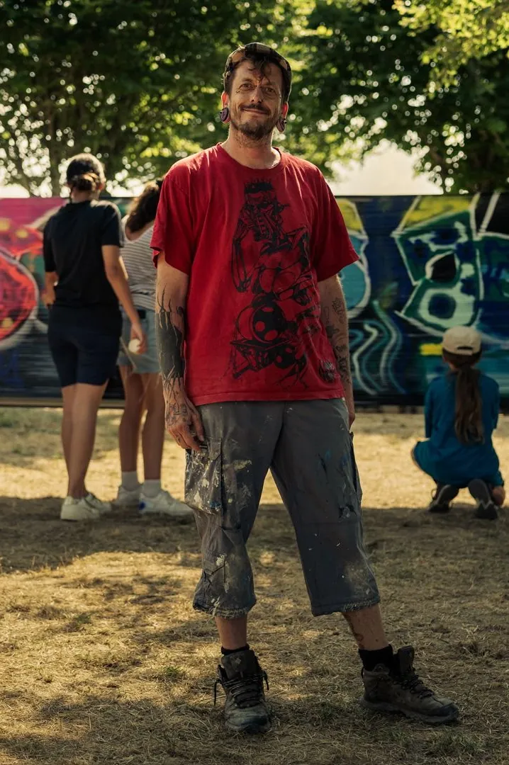 Yannick Jehel debout devant un mur graffé, en tee-shirt rouge et short taché de peinture